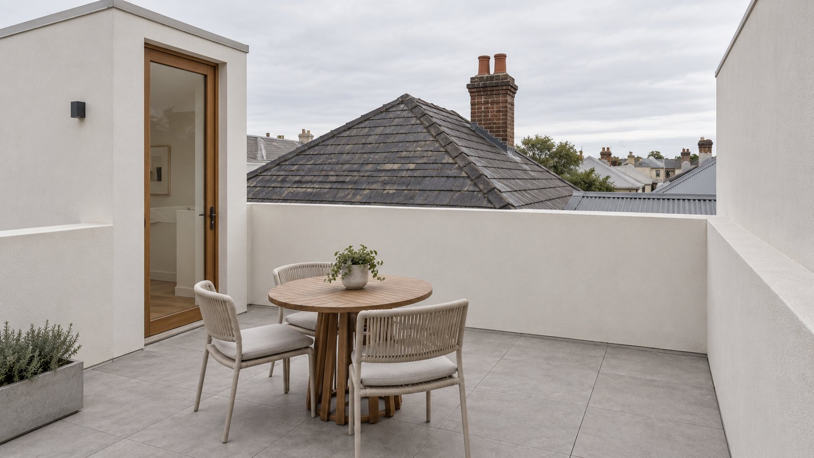 Architectural concept of a small paved roof terrace with solid pale perimeter walls, a timber-framed access door and an older slate roof and brick chimney beyond.