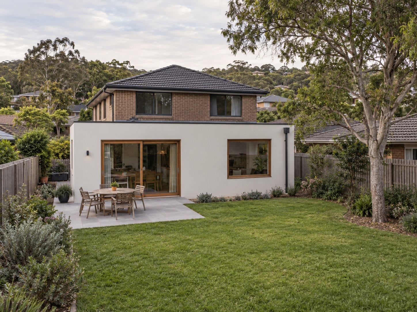 A low white rear room projects beyond a brick upper floor, facing a garden with a small dining terrace, open lawn and established tree.