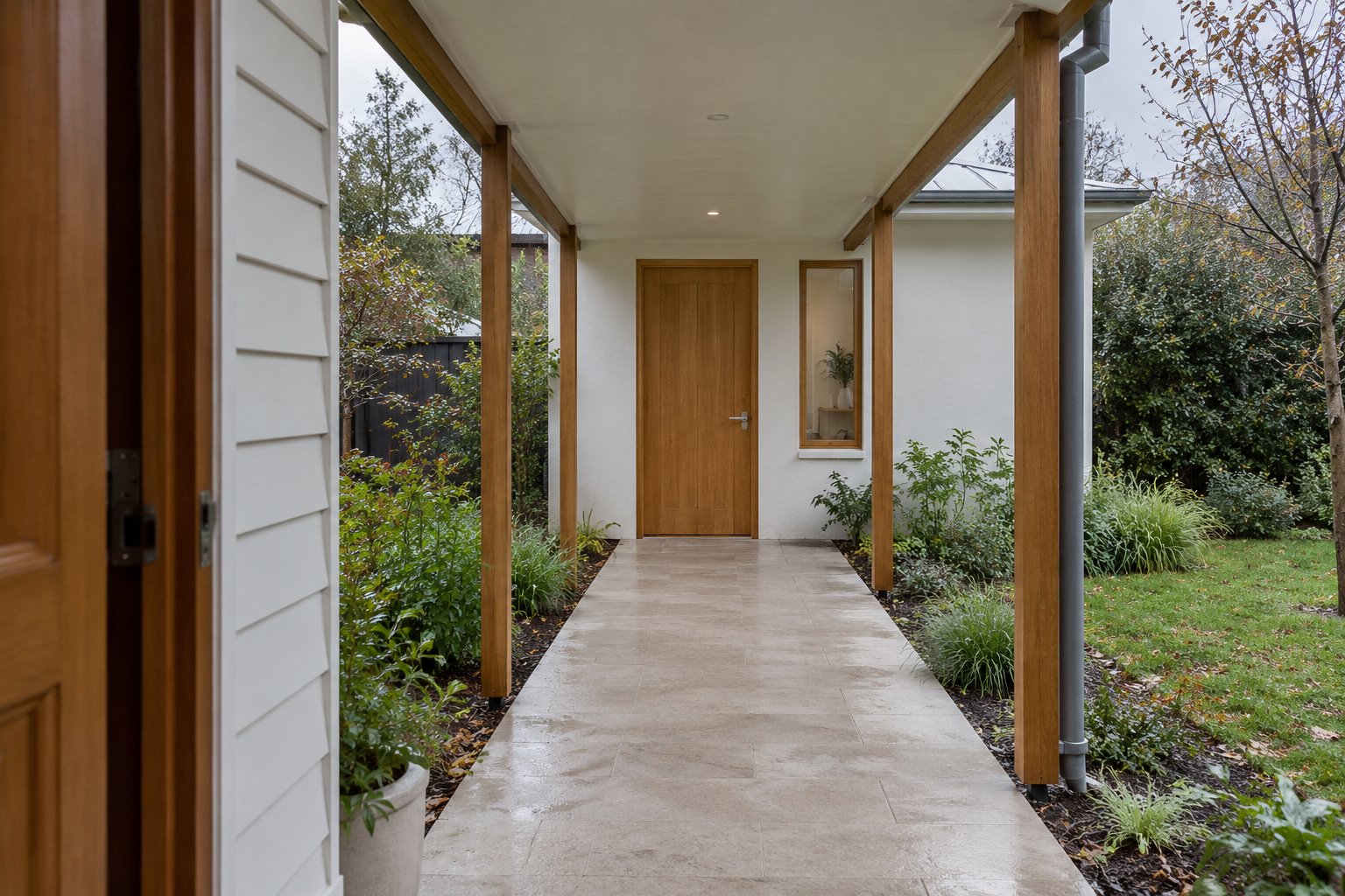 Open-sided covered garden passage leading from a weatherboard house towards a separate room with a closed timber door.