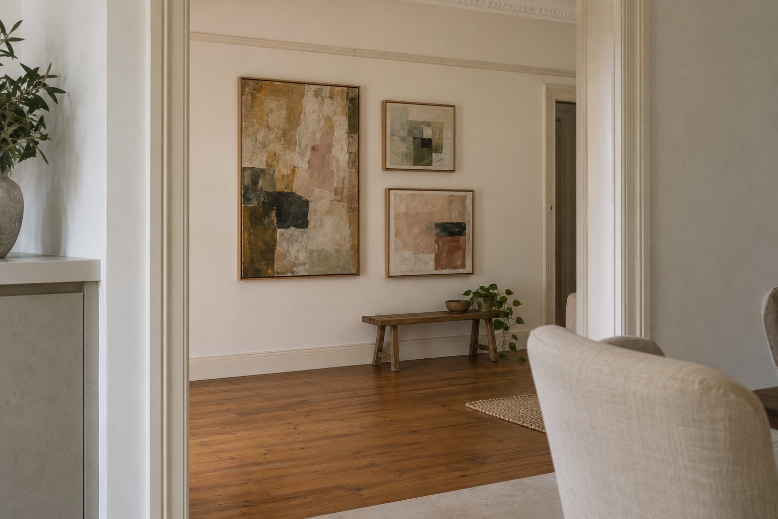 Three differently sized framed abstract pictures on a pale wall beneath a retained picture rail, with a clear viewing area beside a low timber bench.